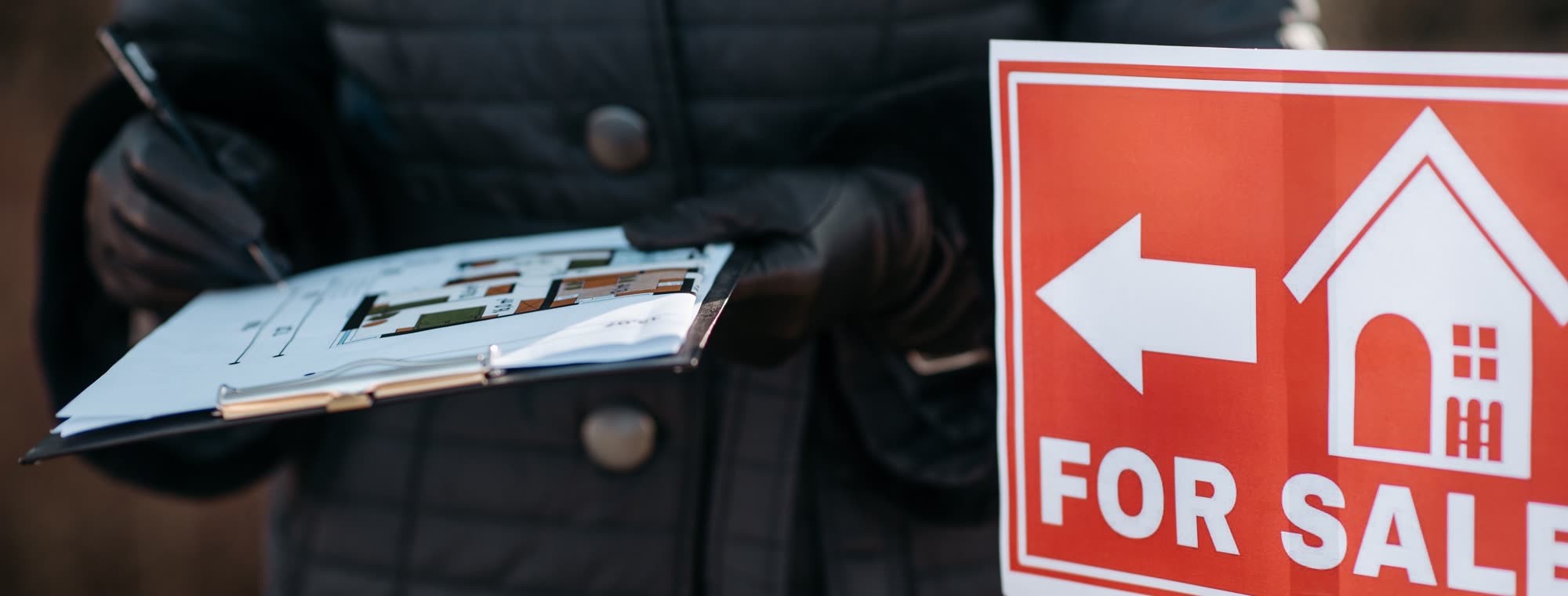 An estate agent holding a floor plan on a clipboard beside a For Sale sign